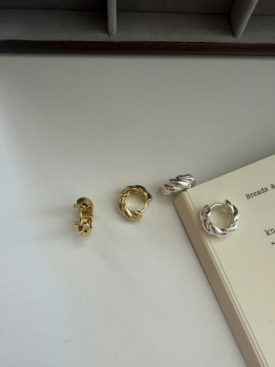 Four twisted metal hoop earrings in gold and silver finishes arranged on a light surface next to an open book.