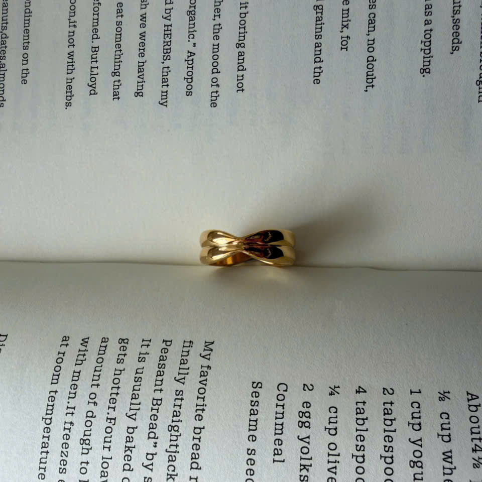 A gold twisted-ring band with black inlay, photographed resting on an open book with printed text visible in the background.